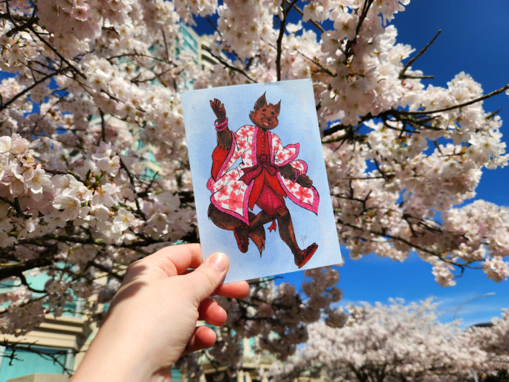 A photo of jam's hand holding a postcard in front of a blooming cherry tree. The postcard has a black catte-person dancing in a Sakura-themed outfit.