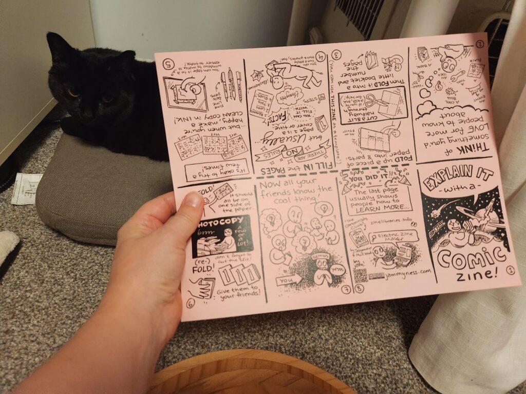 A photo of jam's hand holding the Explain It With A Comic Zine printed on a single sheet of paper. Cat in the background for scale.