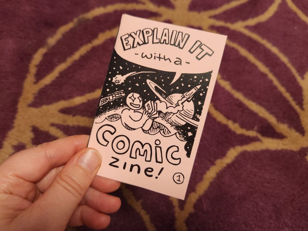 A photo of jam's hand holding a copy of the completed "Explain it with a comic" zine on pink paper